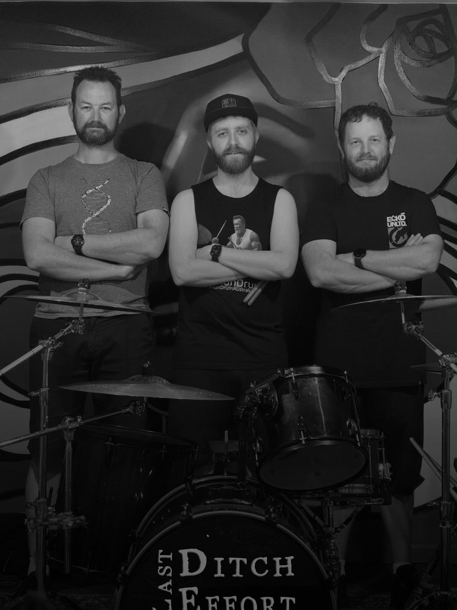 The three members of Last Ditch Effort standing behind a drum kit